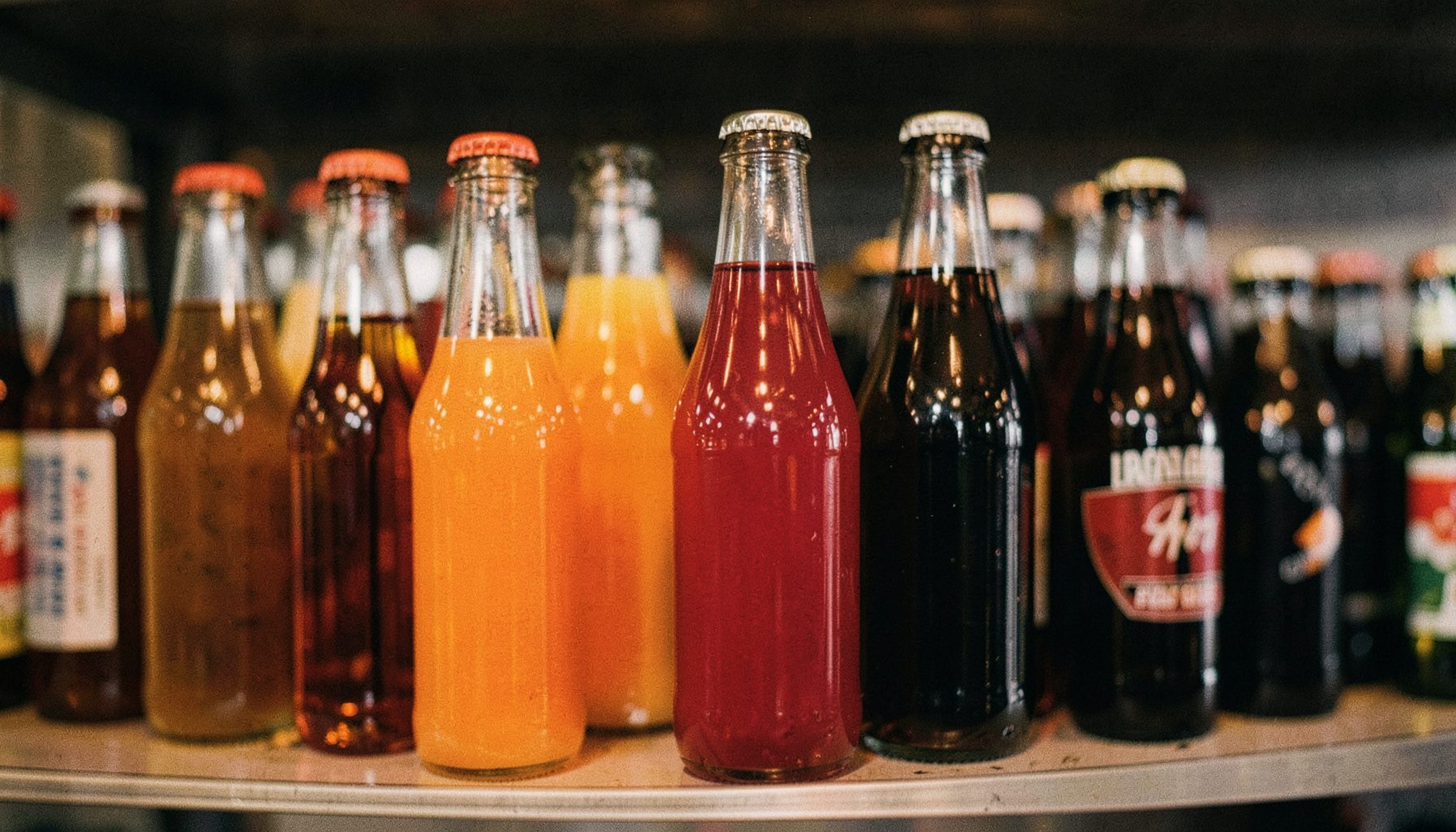 Glass bottles of juice and soda lined up on a shelf, amber and ruby tones