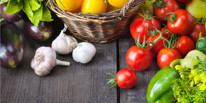 Assortment of fresh vegetables including eggplants, garlic, tomatoes, peppers, and herbs on a wooden table