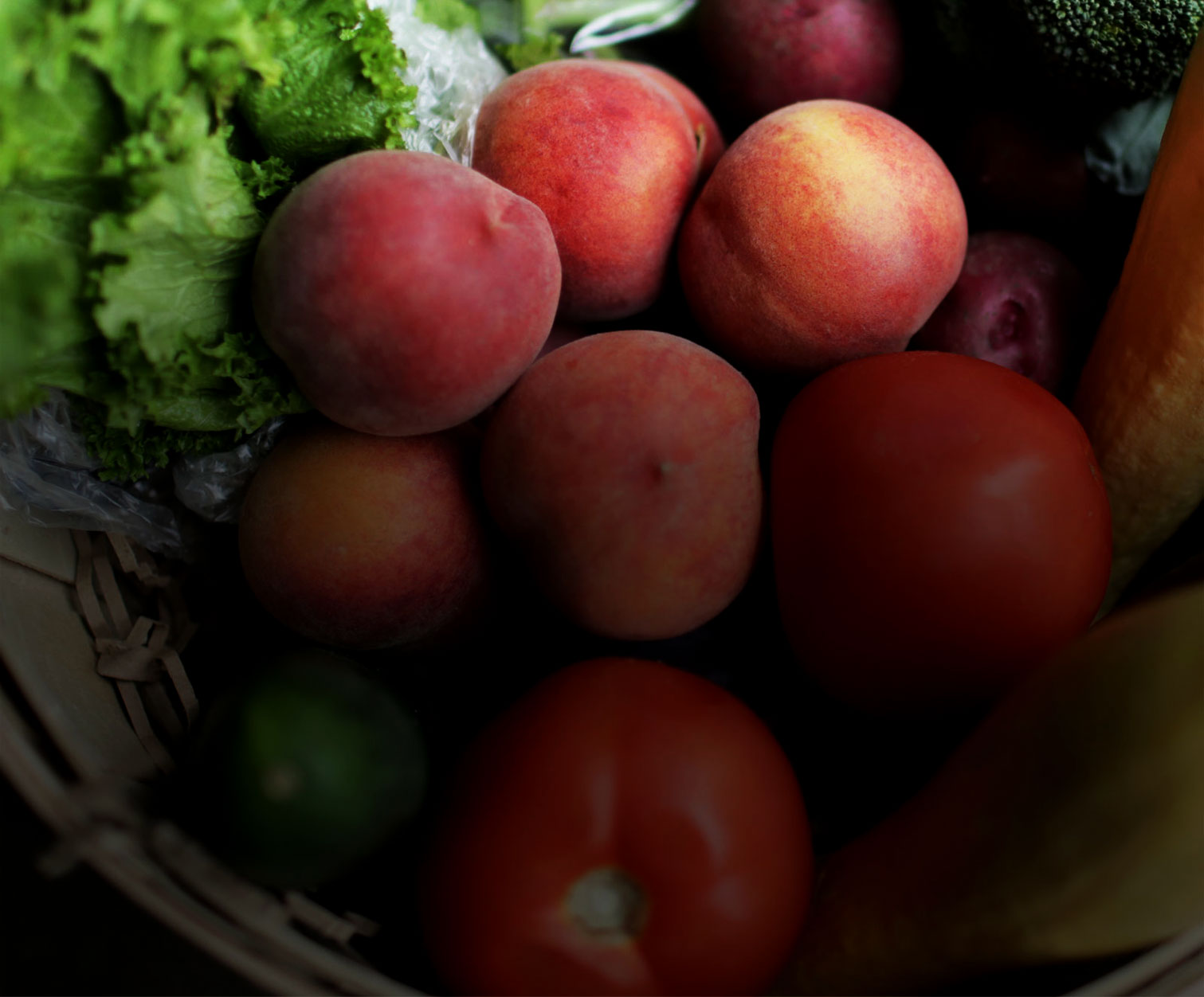 Close-up of fresh produce including peaches, tomatoes, and leafy greens in a basket, warm reddish and green tones, rustic setting